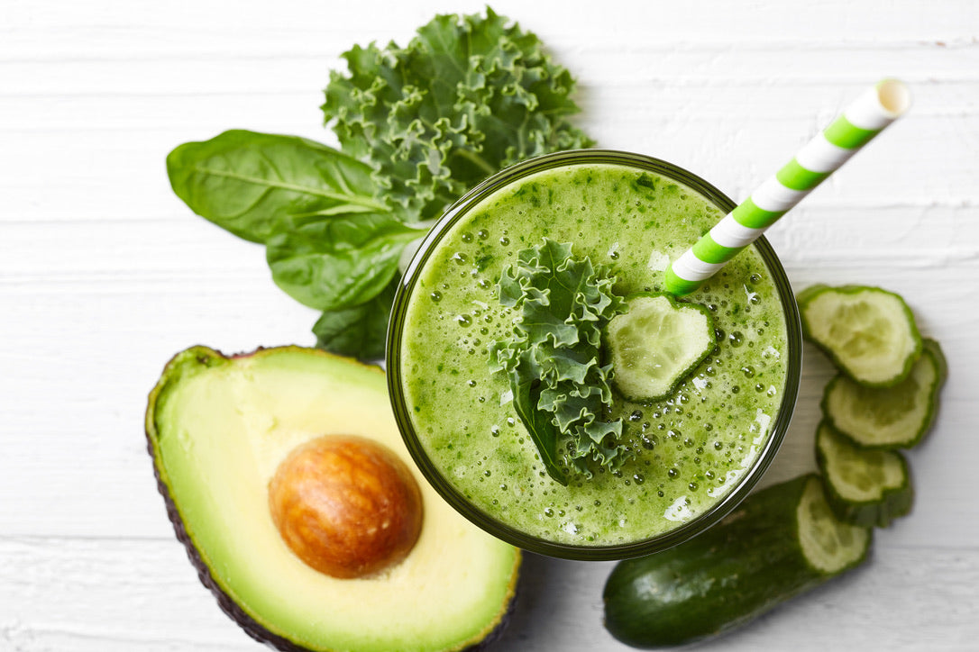 A green smoothie on a board surrounded with cucumbers, salad greens and avocado