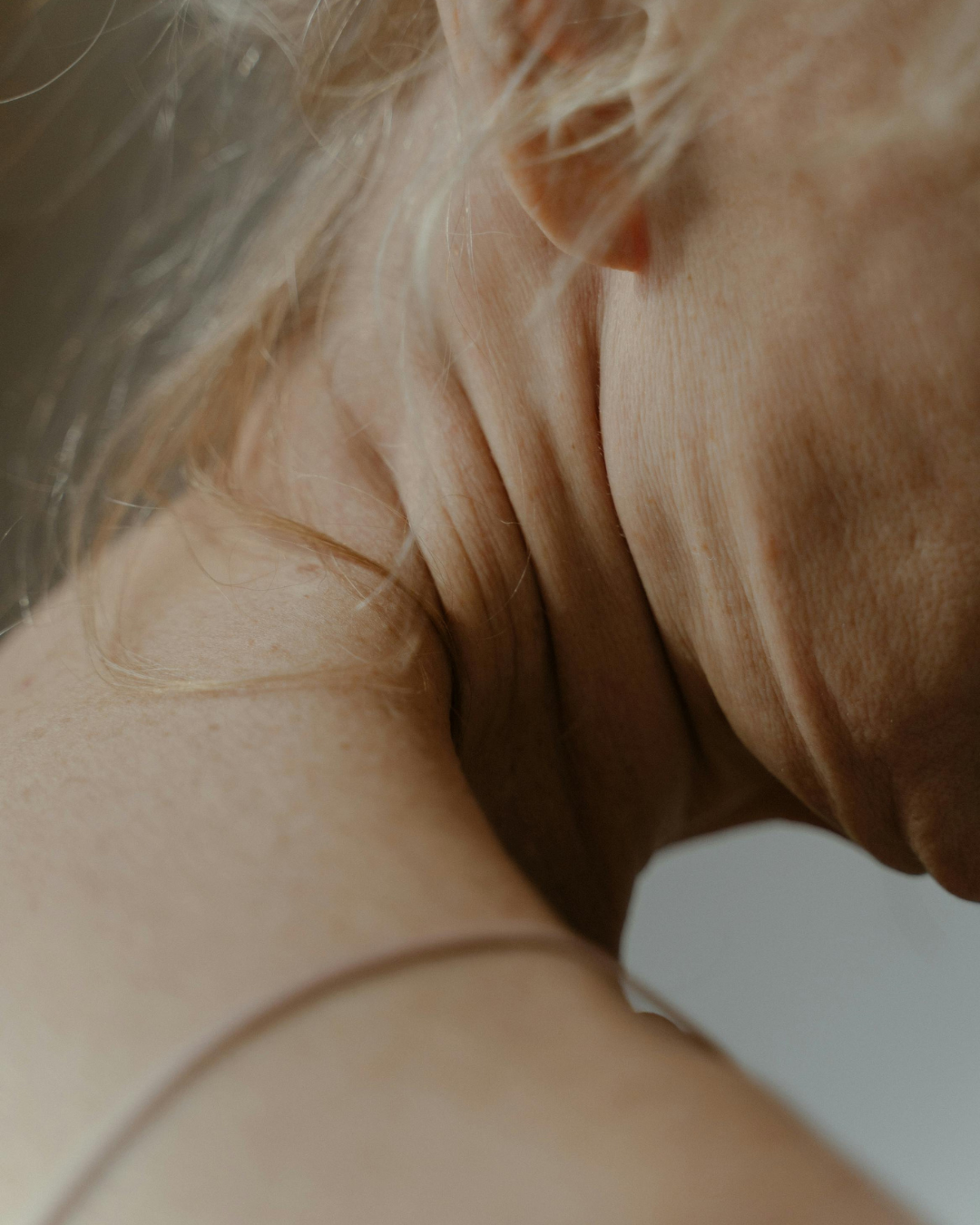 Close-up of a person's neck and shoulder with a thin, delicate cord or strap wrapped around the lower neck, set against a soft neutral background.