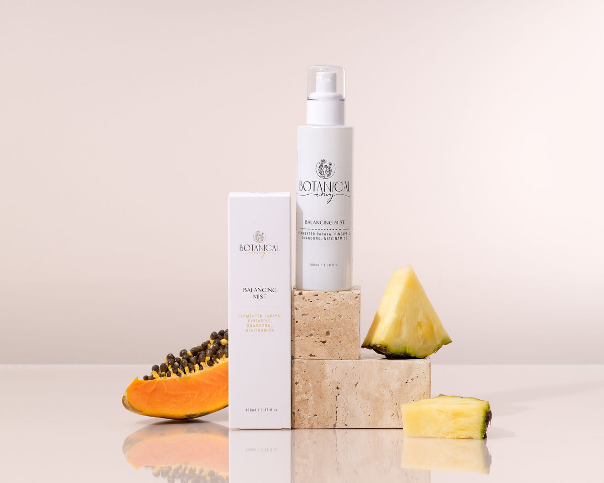 Botanicals Balancing Mist 100ml bottle and matching box displayed on stacked stone blocks, styled with sliced papaya and pineapple wedges on a light beige background.