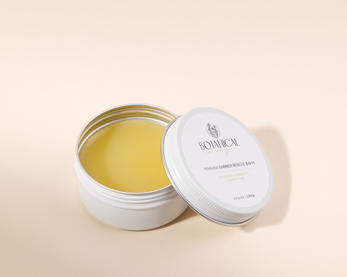 An open round white tin of Botanical Envy manuka Balm with its lid leaning against it, showing the pale yellow balm inside, on a warm beige background.