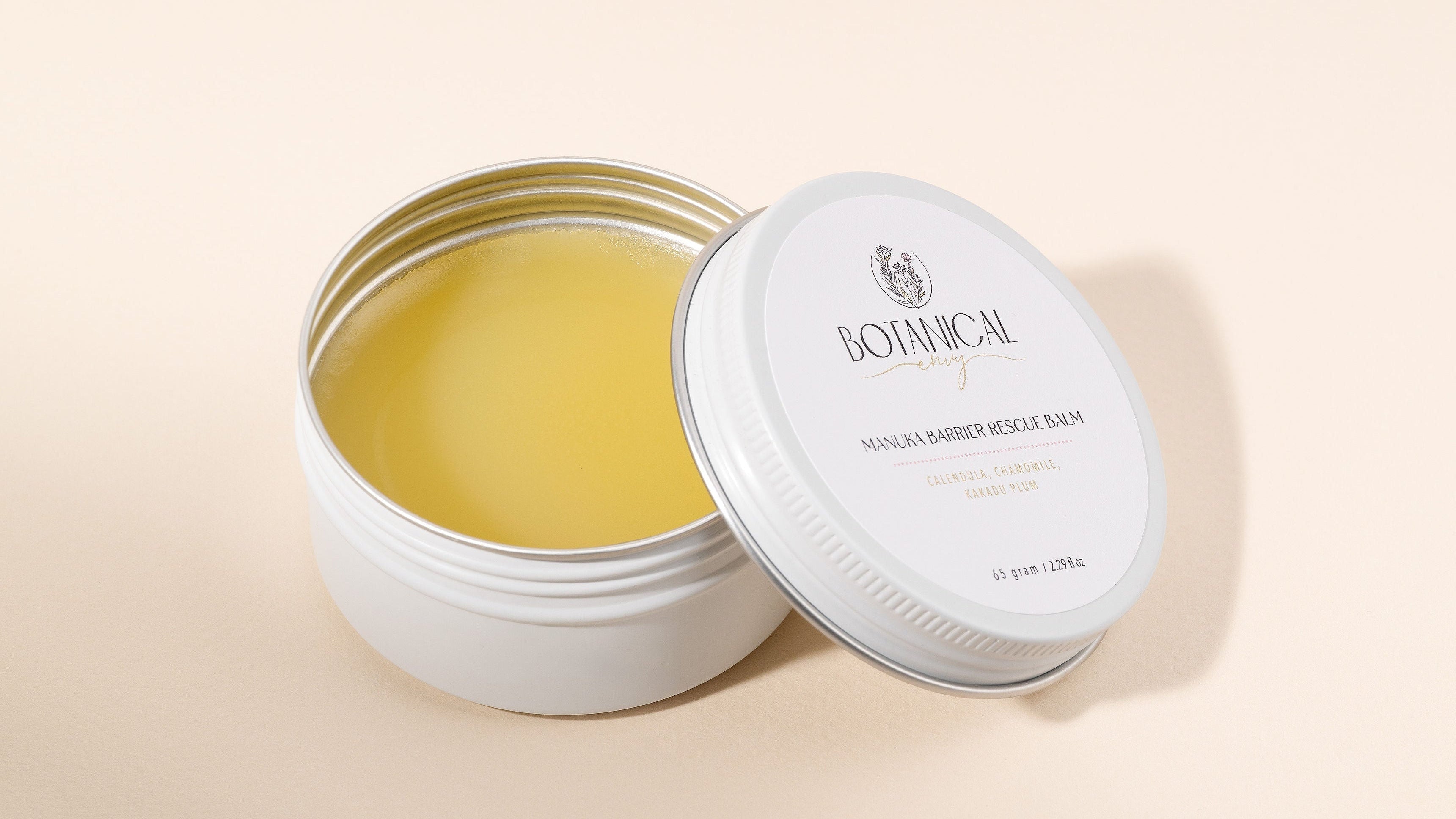 An open round white tin of Botanical Manuka barrier rescue balm with its lid leaning against it, showing the pale yellow balm inside, on a warm beige background.

