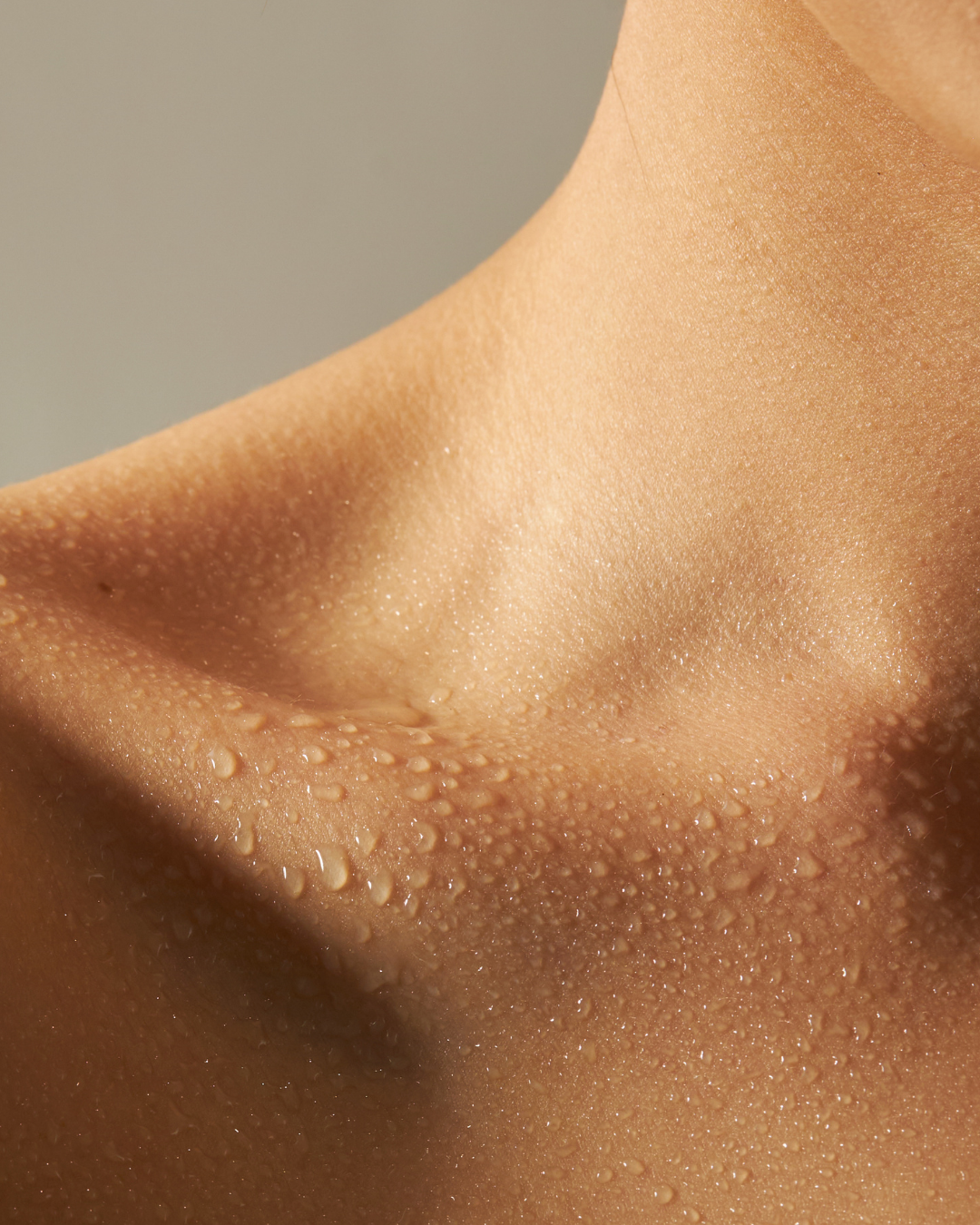 Close-up of a person's neck and upper chest with water droplets on the skin against a soft neutral background.