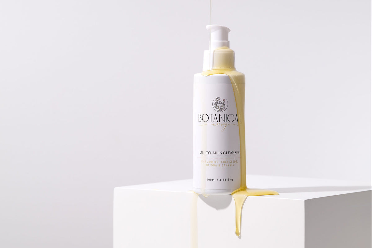 A white bottle of oil to milk cleanser sitting on a white platform with oil dripping from the lid against a white background. 