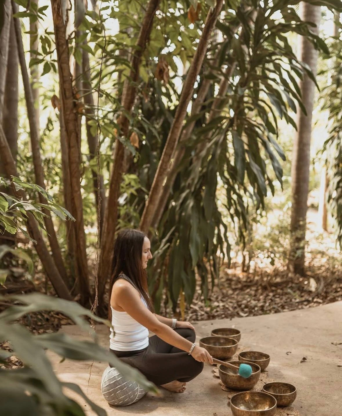 A woman sits cross-legged on a stone path surrounded by dense green foliage, playing a set of metallic singing bowls with wooden mallets.
