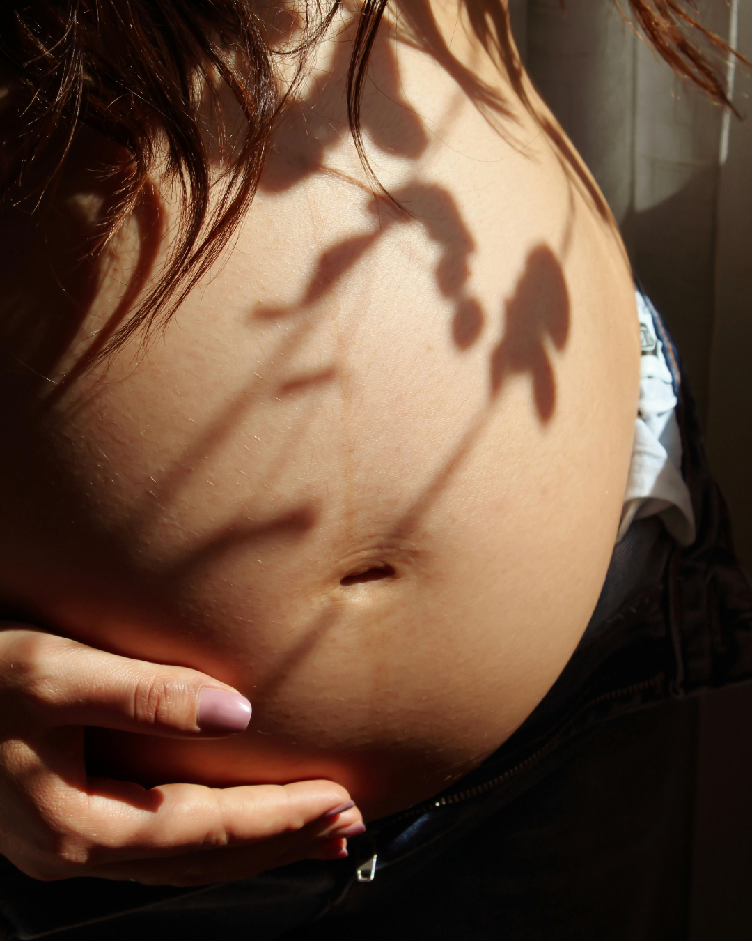 A pregnant lady sitting with a hand on her belly against a window with sunlight and shadow on her. 
