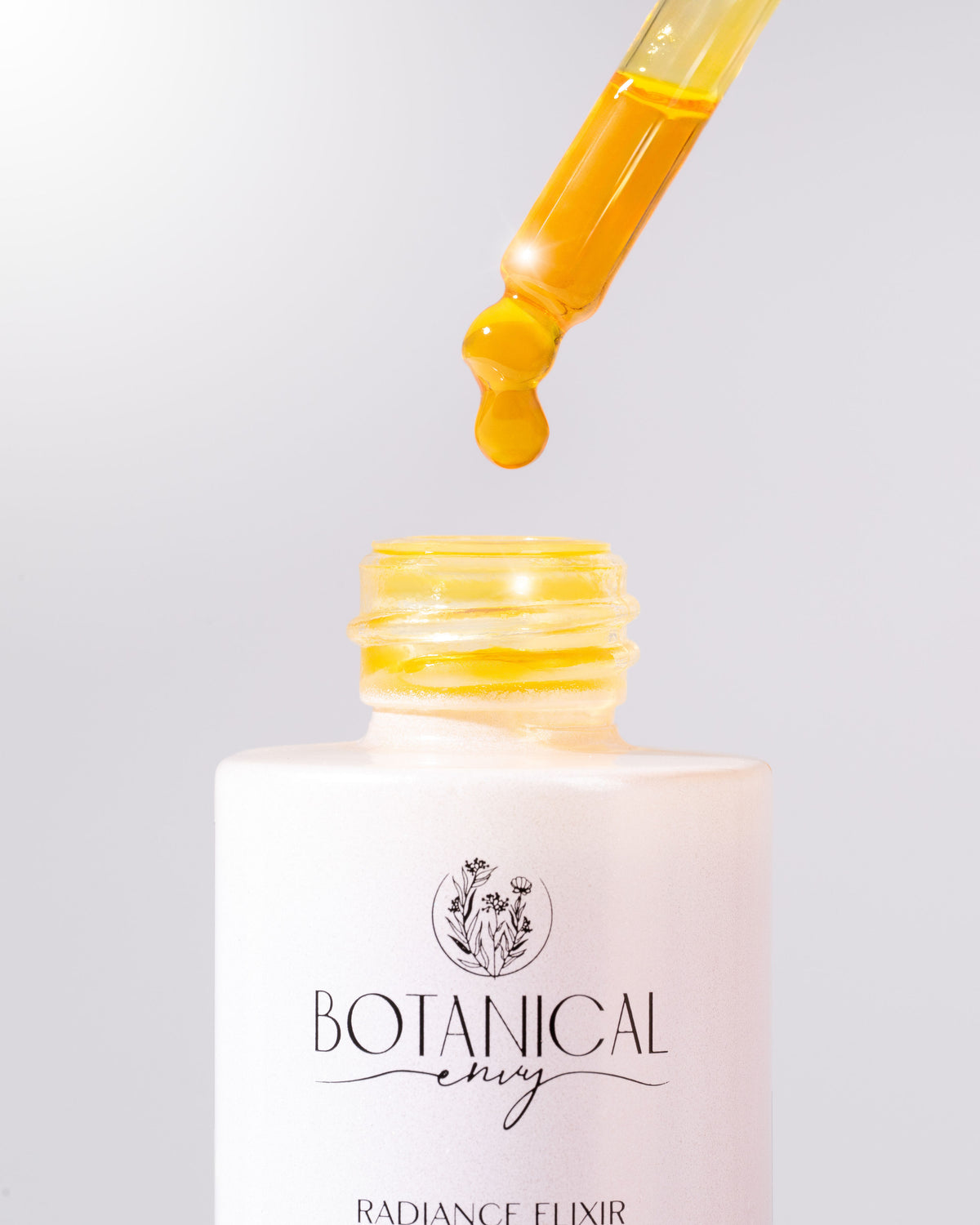 A white bottle of Botanical Envy Radiance elixir with a dropper full of oil against a grey background