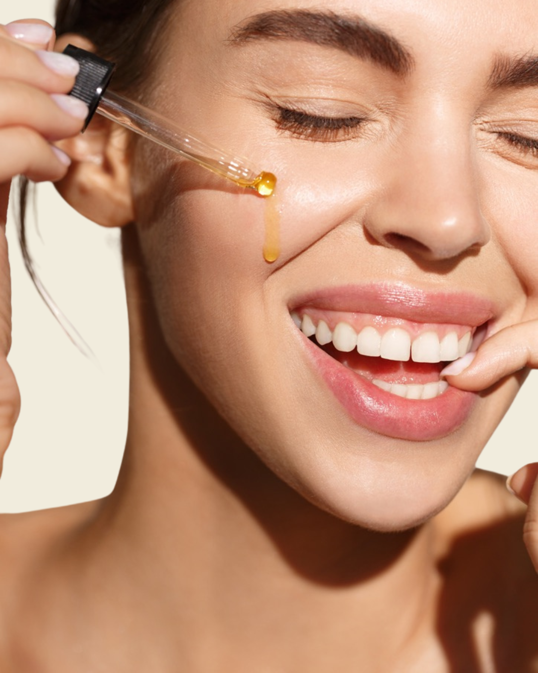 A lady with her eyes closed with a serum dropper full of oil dripping on her face. She is smiling and has a finger in her mouth. 