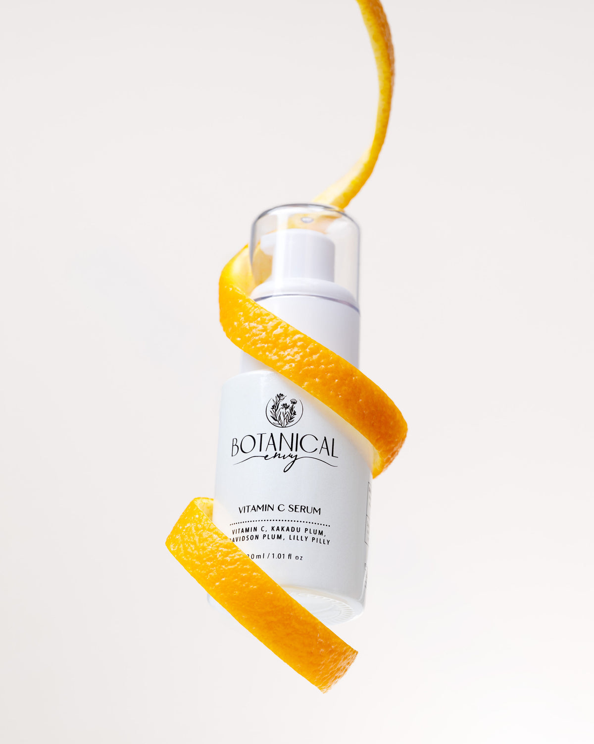 A white glass bottle of vitamin c serum suspended in the air with orange peel wrapped around it against a light beige background.