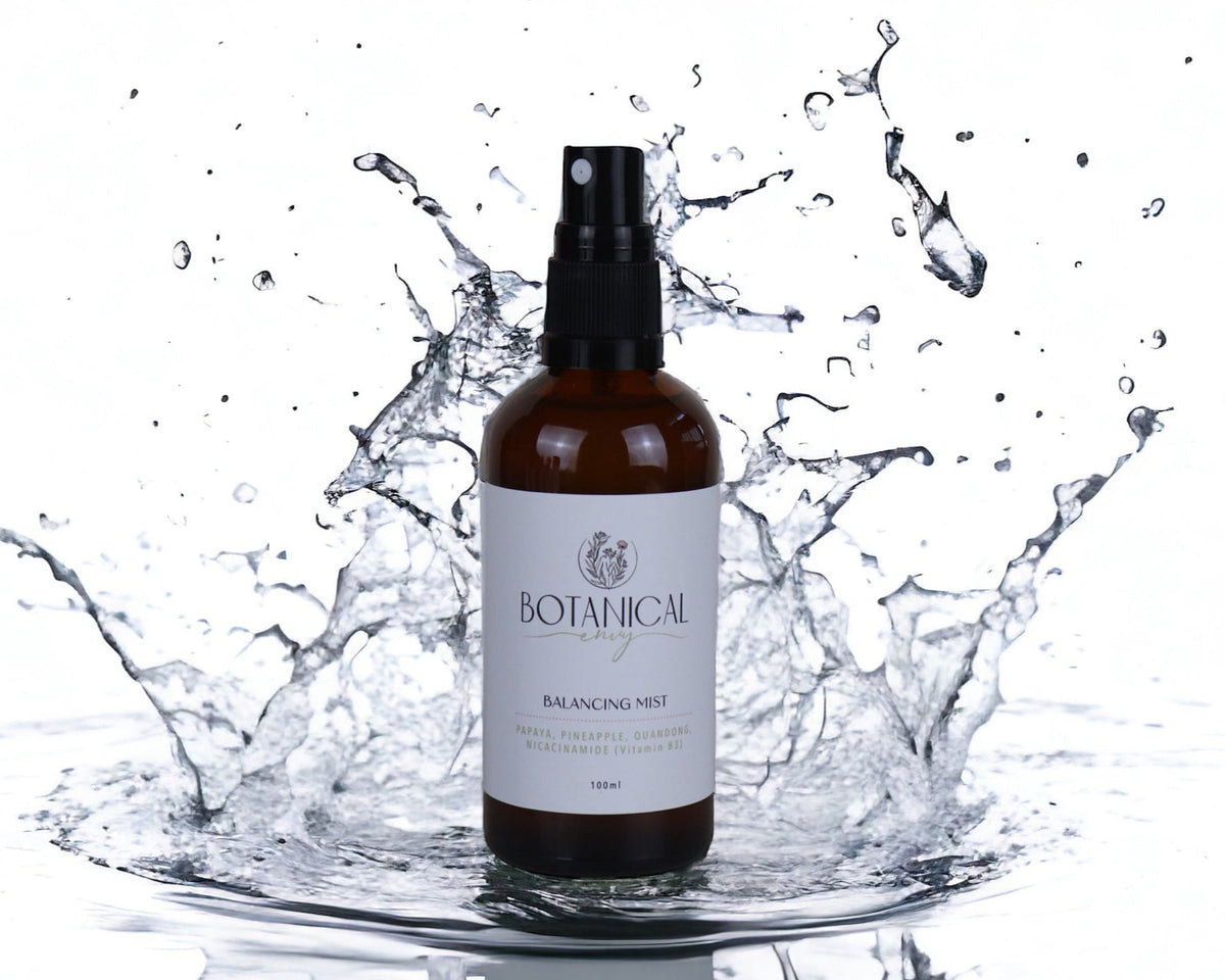 A brown glass spray bottle labeled 'Botanicals Balancing Mist' stands in a water splash on a white background, with droplets frozen around it.